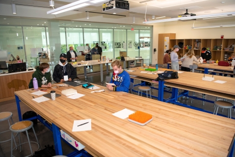 Students in classroom around large wooden desk. 
