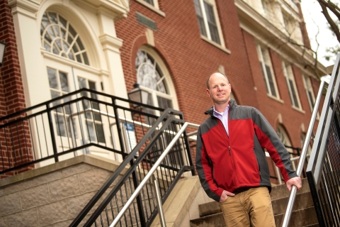 Eric Kennedy wears a red and gray fleece jacket and stands and smiles on the steps outside a building.