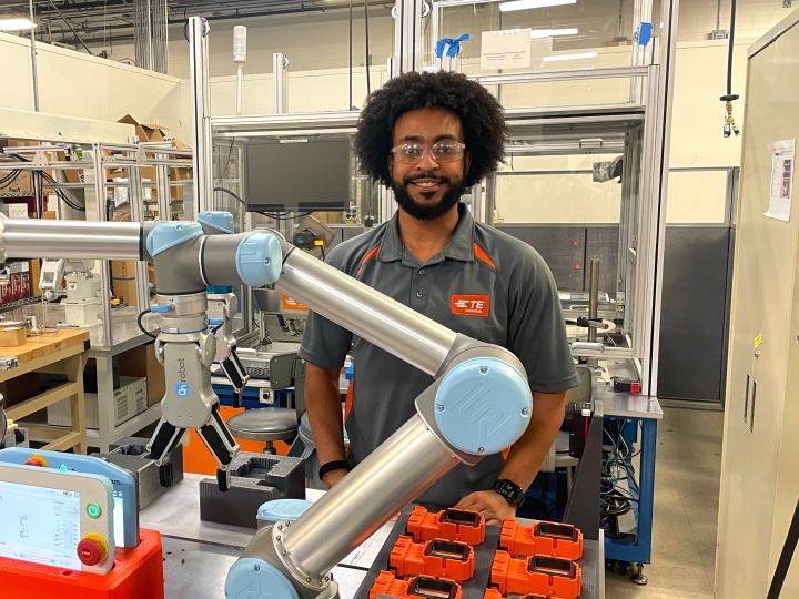 Kellen Haile stands in a factory-like setting wearing safety goggles and standing behind a large robotic arm.