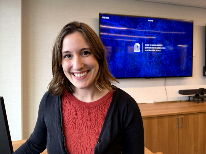 Juliet King smiles in a room where a large screen is on the wall behind her with the University of North Carolina's logo