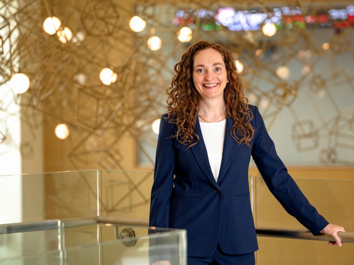 Portrait of a woman wearing a blue suit with her hand on a railing in a bright lobby with a lighted hanging sculpture behind her.