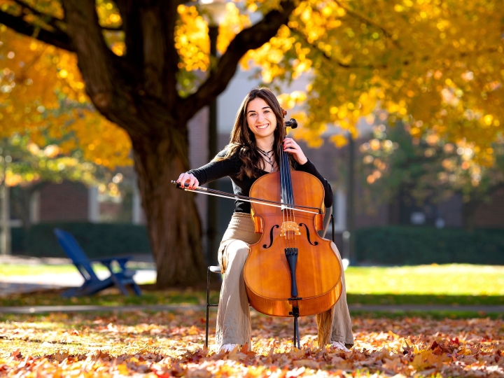 Elle Chrampanis sits on a chair and plays the cello while sitting outside under a tree with golden leaves that hang from the branches and also sit on the ground around her..