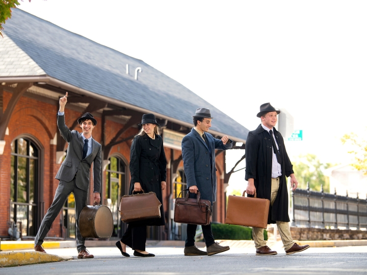 Four people dressed as salesmen cross an intersection in front of a trainstation.