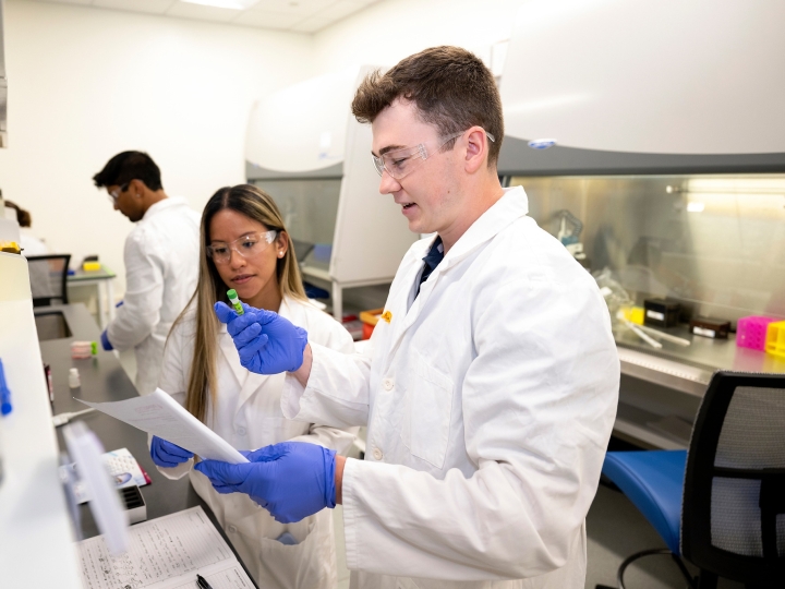 Professor Olivia Boerman and Connor Kozick &#039;26 are wearing white lab coats and purple gloves in a lab setting.