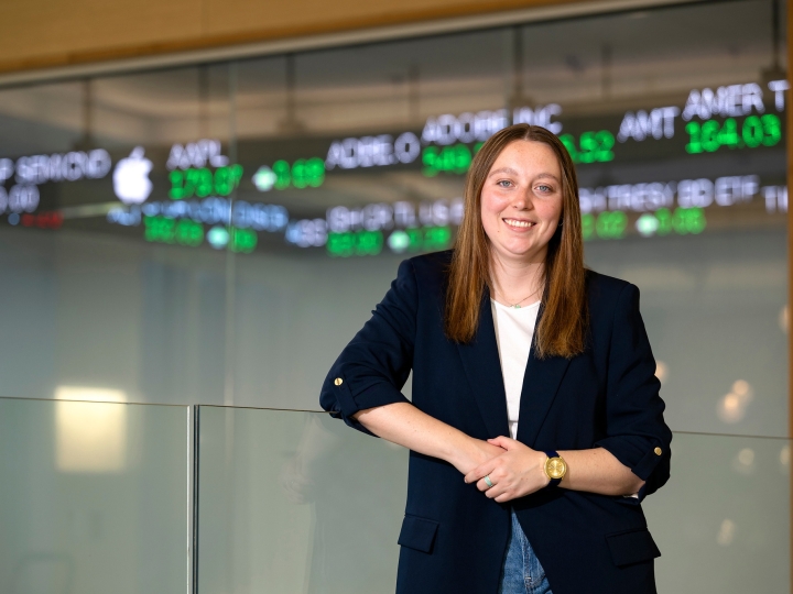 Caitlin Yant &#039;24 stands and smiles in the Moriatry Investment Center in Holmes Hall with a stock ticker behind her.