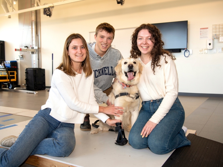Three students sit and smile with a golden retriever on the ground inside a work space. The dog wears a prosthetic on its front right leg.