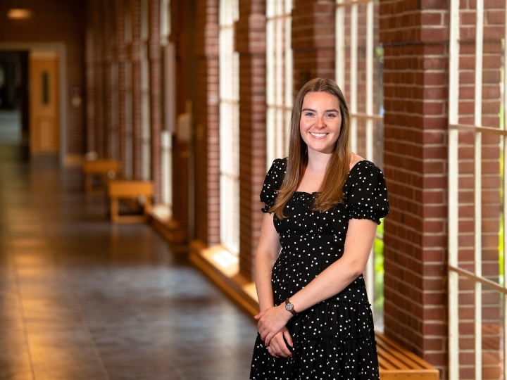 Kendall Robertson wears a black dress and smiles while standing in a hallway with brick columns and windows behind her.