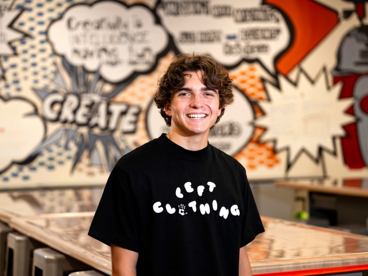 Mikey Brandt wears a black Tshirt with a LEFT Clothing logo printed across his chest. He is in a Makerspace with a graffiti-style wall design behind him.