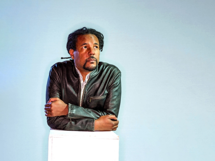 Portrait of author Colson Whitehead leaning forward against a white box, staring upward and off camera to the right.