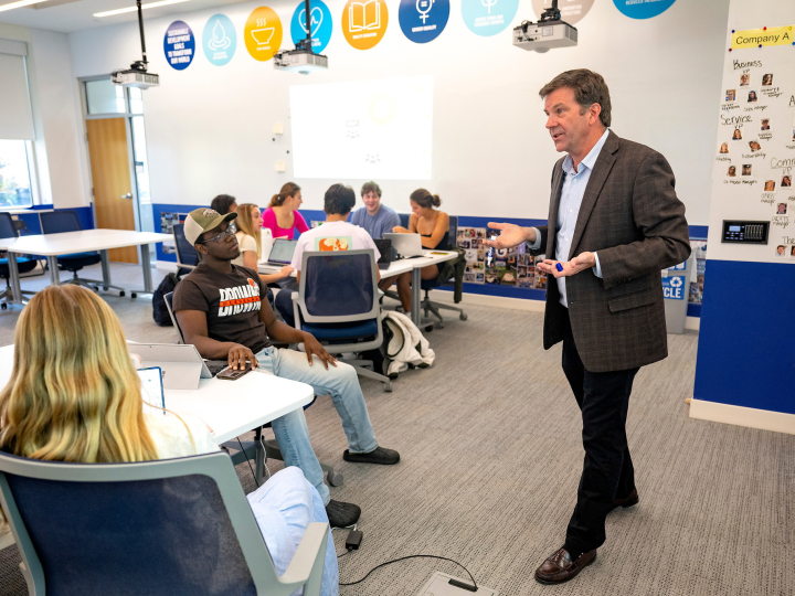 A professor in a suit jacket stands in front of a white board with students surrounding him in a classroom.