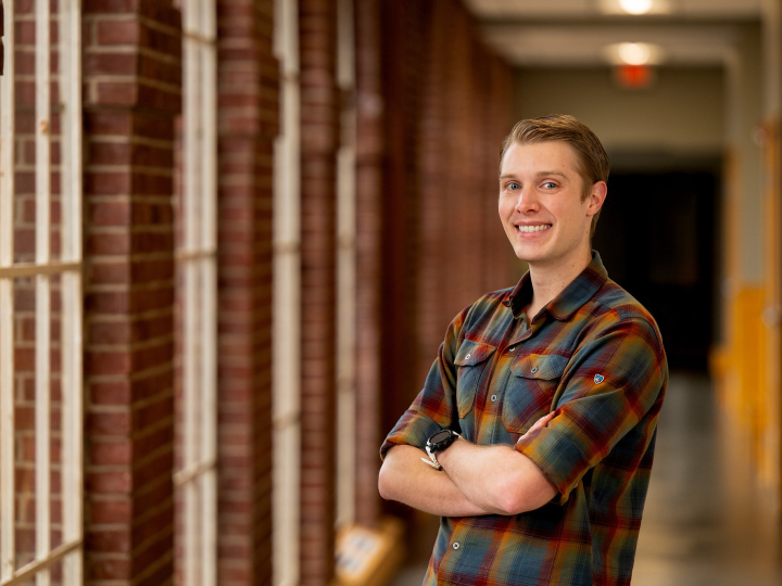 Chris Gregory wears a plaid shirt and stands in a hallway with window and brick trim.
