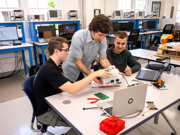 Two students sit at a table in a makerspace with a staff member in between them as they look at pieces of technical hardware together.