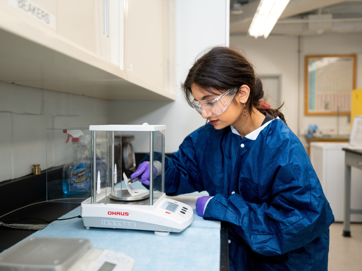 Priyanka Poplai '29 wears a blue lab coat, blue gloves, and safety goggles as she works in a lab space.