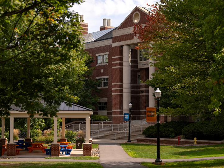 a scenic shot of campus features a covered patio with orange blue chairs, with Rooke Science Center in the backdrop and lush green foliage.