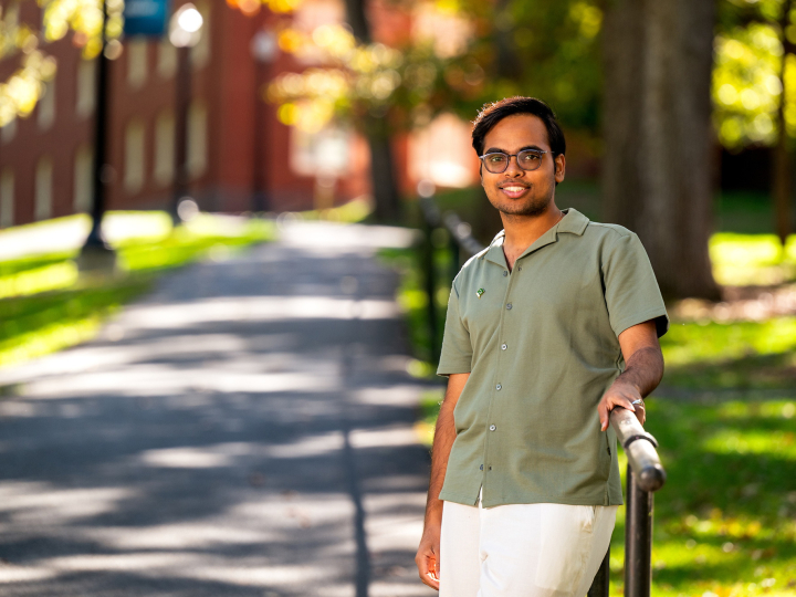 A Bucknell student wearing a green shirt stands outside on a sunny day with trees and a walking path in the background.