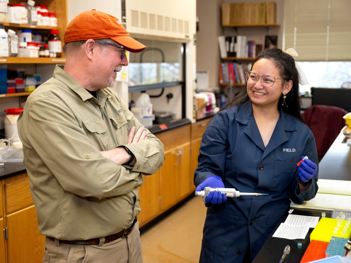 Angel Eunisse Yanga is smiling in a campus lab space wearing a blue lab coat while talking with Professor Ken Field.