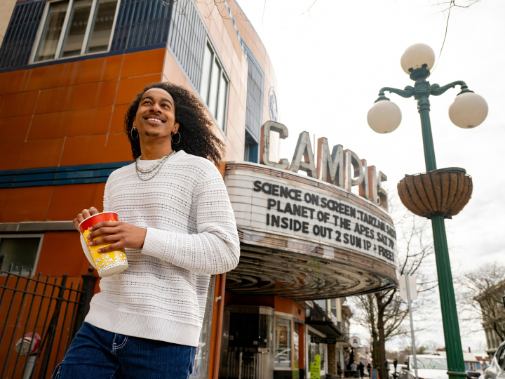 Caleb Warren smiles as he holds a small container of movie theatre popcorn; the marquee of the Campus Theatre is behind him.