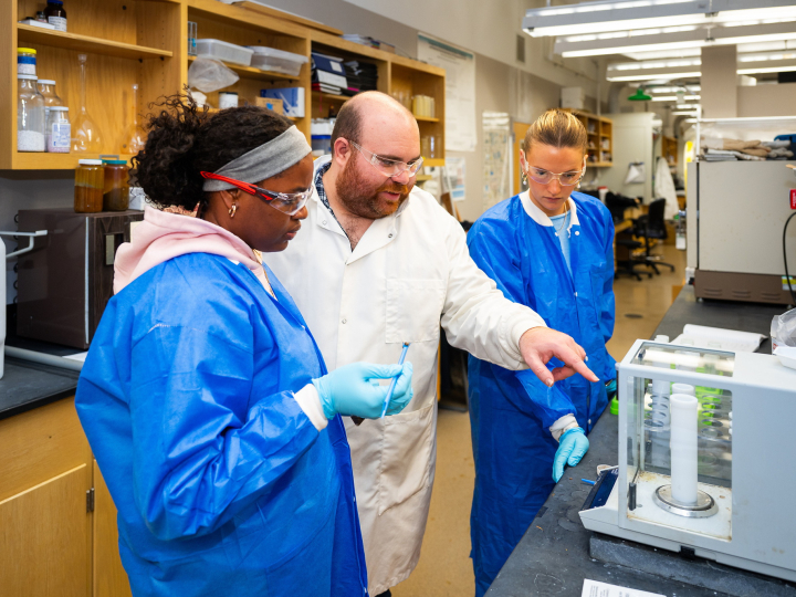 Two students wearing blue lab coats and a professor in a white lab coat stand inside of a laboratory.