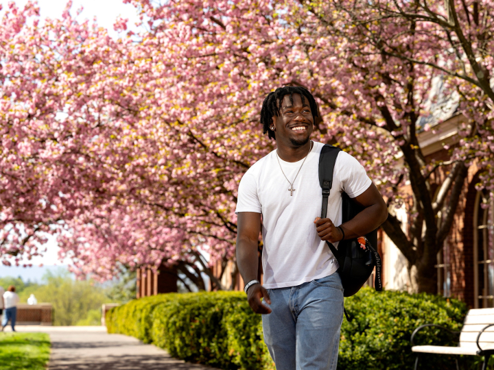 Momo Peters wears a white T-shirt and jeans with a backpack slung over his left shoulder as he smiles and walks on campus with pink cherry blossoms behind him.