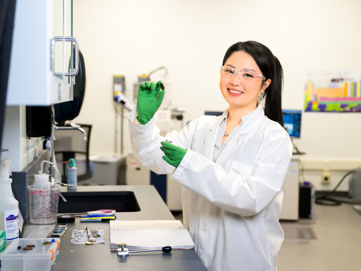 My Ly wears a white lab coat, safety goggles and green gloves while holding lab equipment in a lab setting.