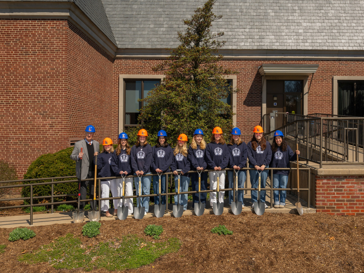 a group of students wearing orange and blue hard hats and blue hooded sweatshirts stand in a line together with President John Bravman outside the ELC.