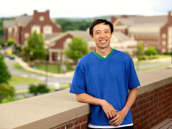 Sing Chun Lee wears a bright blue shirt and leans against a brick wall while smiling on campus