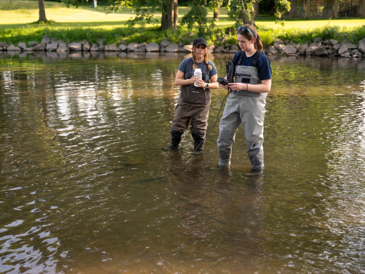 Two researchers wearing chest waders stand in a shallow waterway, one holding a sample bottle and the other checking a handheld device.