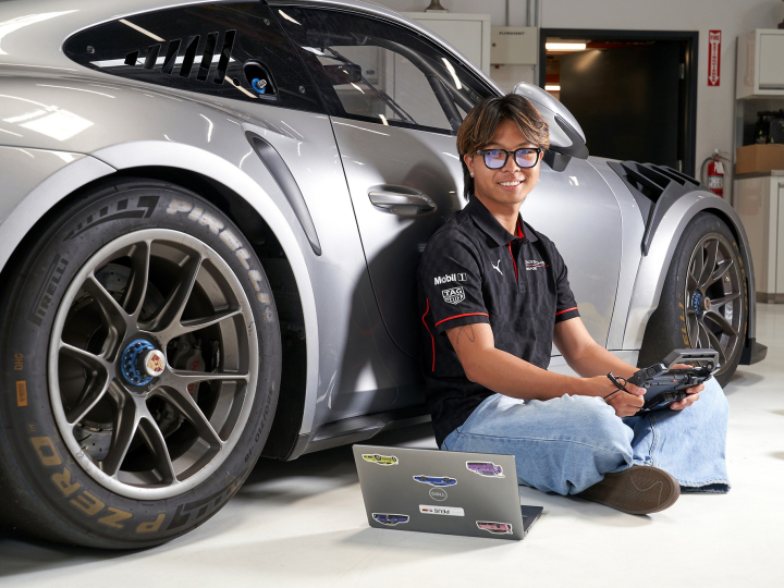Zach Ebiane sits on the ground with a laptop while leaning against a silver Porsche.