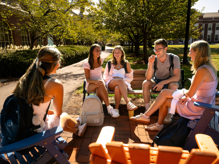 A group of students sits outside on benches in the sunshine. They are smiling.