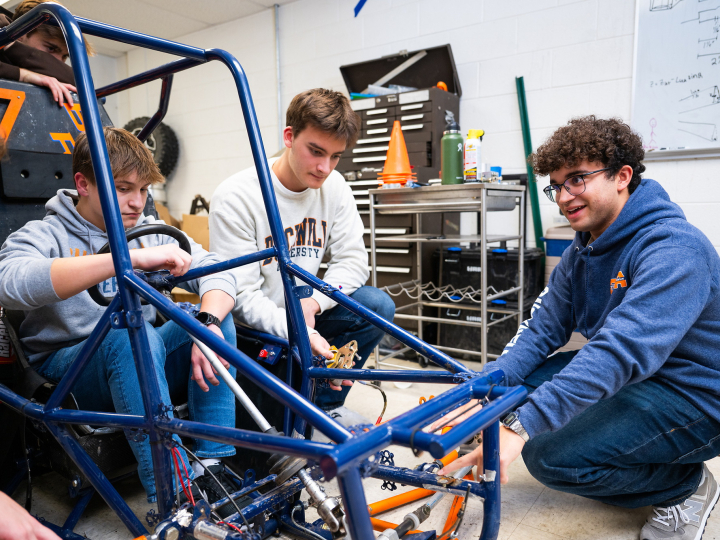A group of students gather around the frame of a stripped down vehicle.