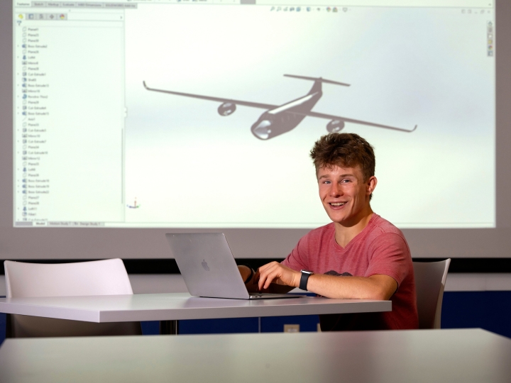 Kade Davidheiser sits at a table on his laptop with an image of an airplane model projected on the screen behind him