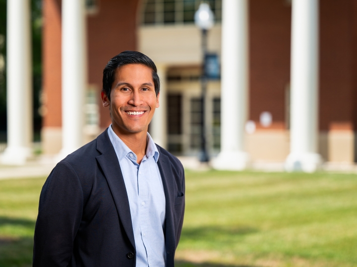 Professor Karlo Malaga stands on campus and smiles, a building with columns is in the background