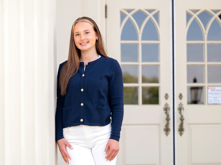 Madelyn Hudak wears a blue top and white pants and leans slightly against a white column outside a building on campus.