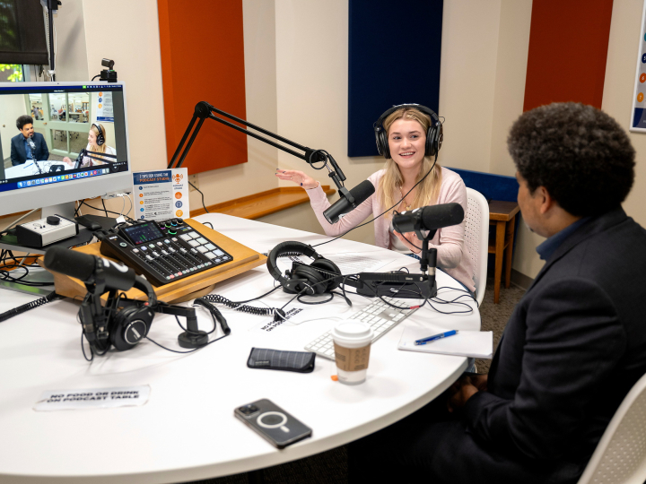 Rayna Tyler '27 wears headphones and sits at a table with a microphone and computer screen while speaking to a guest, who has his back to the camera.