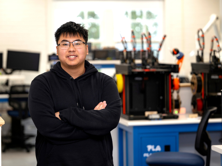 Richard Chi stands in a makerspace and smiles while wearing glasses and a navy hoodie.