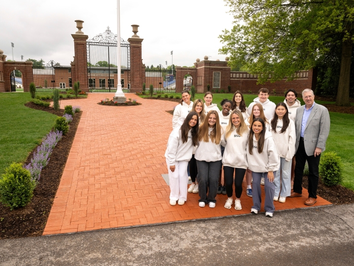A group of students stands with President John Bravman at the bottom of a wide, brick pathway that leads through the Christy Mathewson Gates on campus.