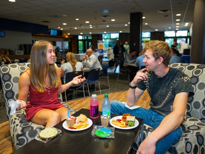Two students sit in The Bison on colorful chairs and talk while eating food.