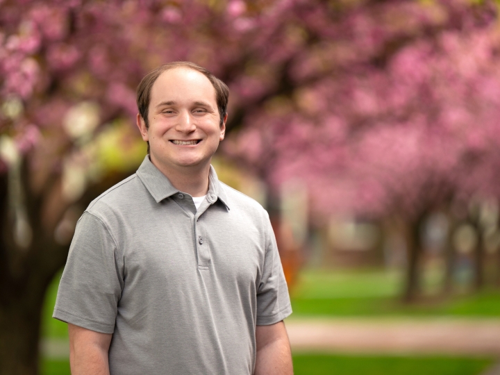 Zane Hensal wears a gray short-sleeved colored shirt and smiles on campus with cherry-blossom trees behind him.