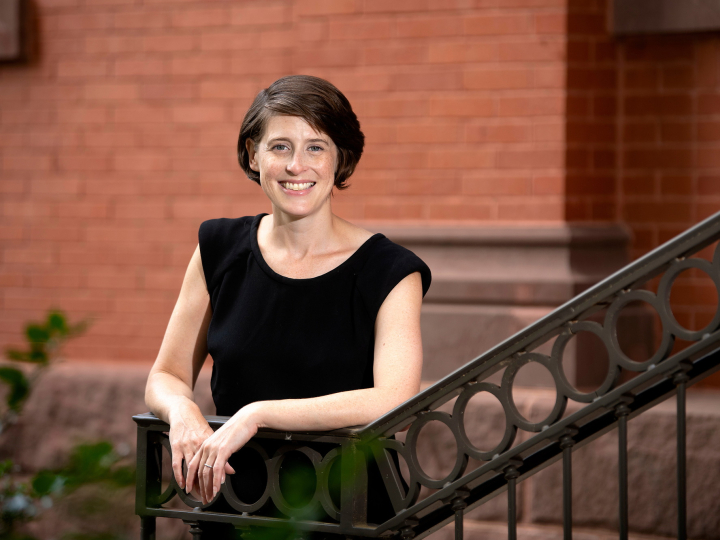 Reggie Gazes wears a black sleeveless shirt and stands against the railing of a staircase outdoors on campus with a red brick building behind her.