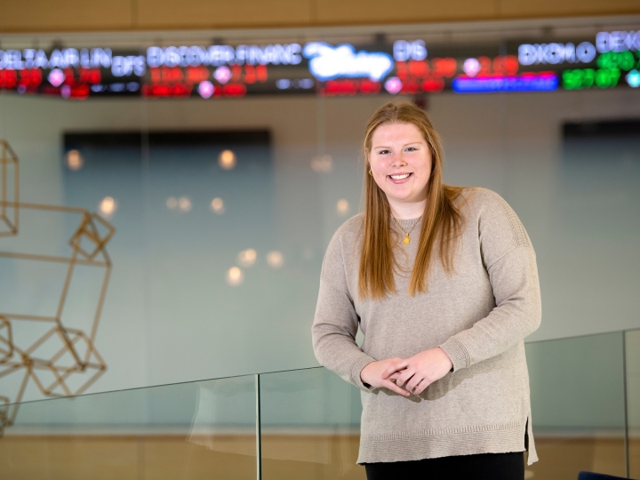 A student stands inside a building with a high-tech ticker behind her, displaying current stock prices. 