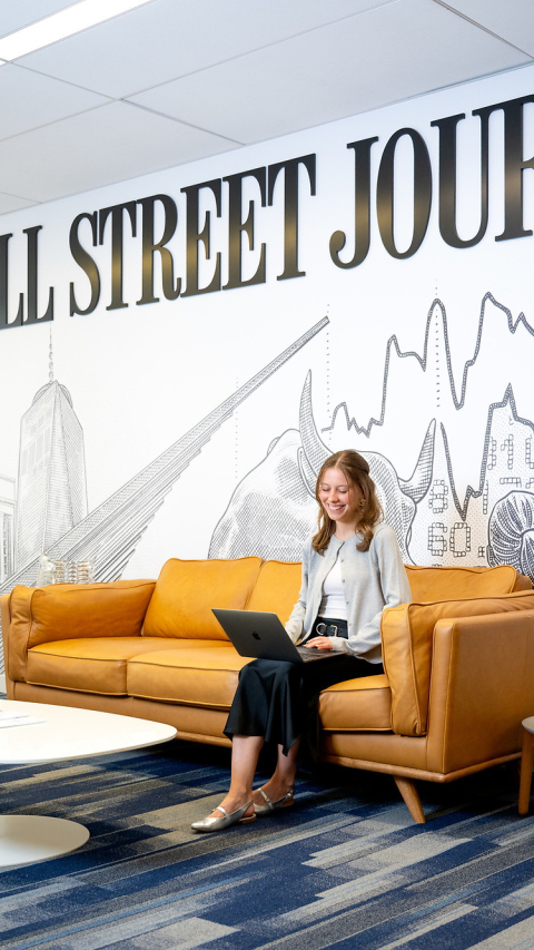 Student sitting on an orange couch with her laptop, under a sign that says Wall Street Journal