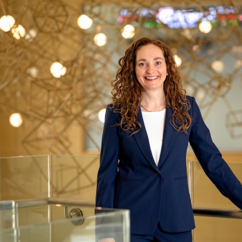 Portrait of a woman wearing a blue suit with her hand on a railing in a bright lobby with a lighted hanging sculpture behind her.