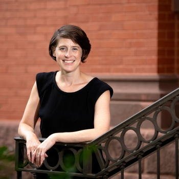Reggie Gazes wears a black sleeveless shirt and stands against the railing of a staircase outdoors on campus with a red brick building behind her.