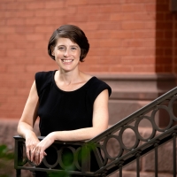 Reggie Gazes wears a black sleeveless shirt and stands against the railing of a staircase outdoors on campus with a red brick building behind her.