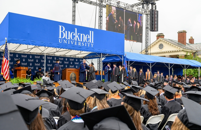 Graduates sit in front of the Bucknell Commencement platform in caps and gowns while students walk across stage to receive their diplomas.