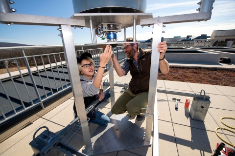 A female student is looking under a metal frame at a mechanism that drives a vertical wind turbine. A male professor kneels across from her.