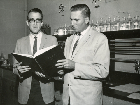 A vintage photograph shows two men in lab coats examining a leather-bound book in a chemistry lab.