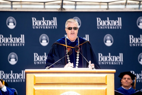 Bucknell president John C. Bravman stands at a podium on the Commencement stage, smiling and wearing sunglasses.