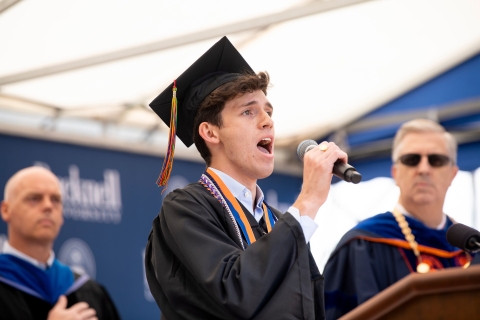 Tom McKillop ’23, dressed in a black graduate's gown and mortarboard, sings the national anthem into a microphone
