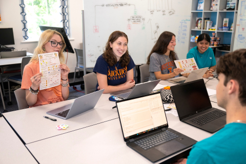 Five students sitting at a table with their computers. one student is holding up a piece of paper, another is smiling. 